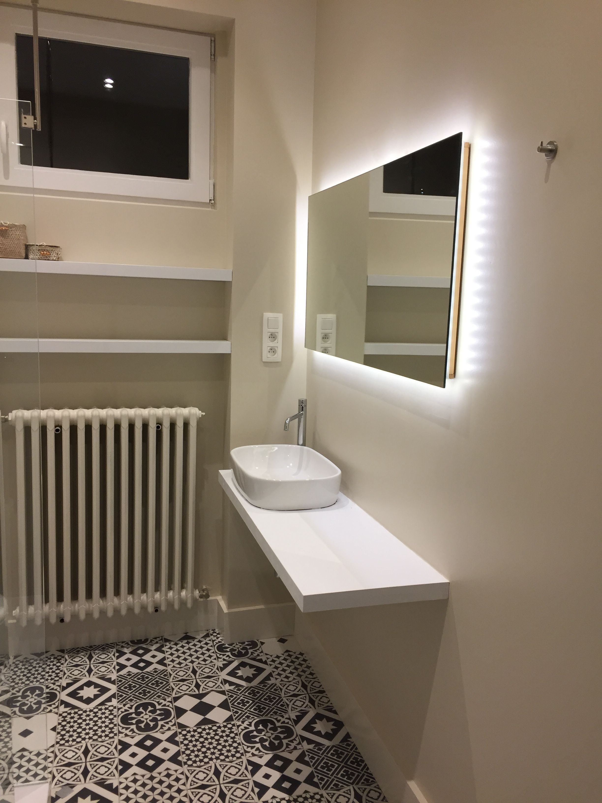 Une salle de bain moderne avec un lavabo blanc, un miroir doté d’un éclairage indirect, une fenêtre et des carreaux de sol à motifs.