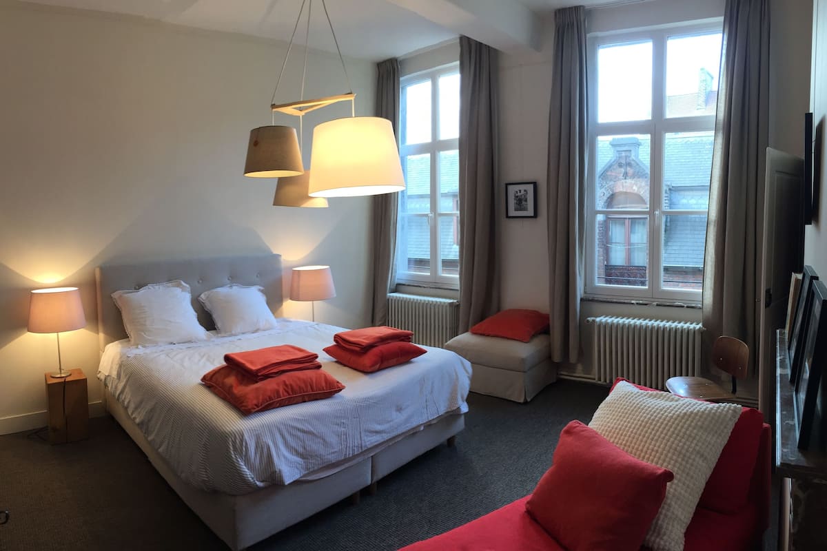 Ein Hotelzimmer mit einem großen Bett, zwei Nachttischlampen, einem roten Sofa und einem Fenster mit Blick auf Gebäude.