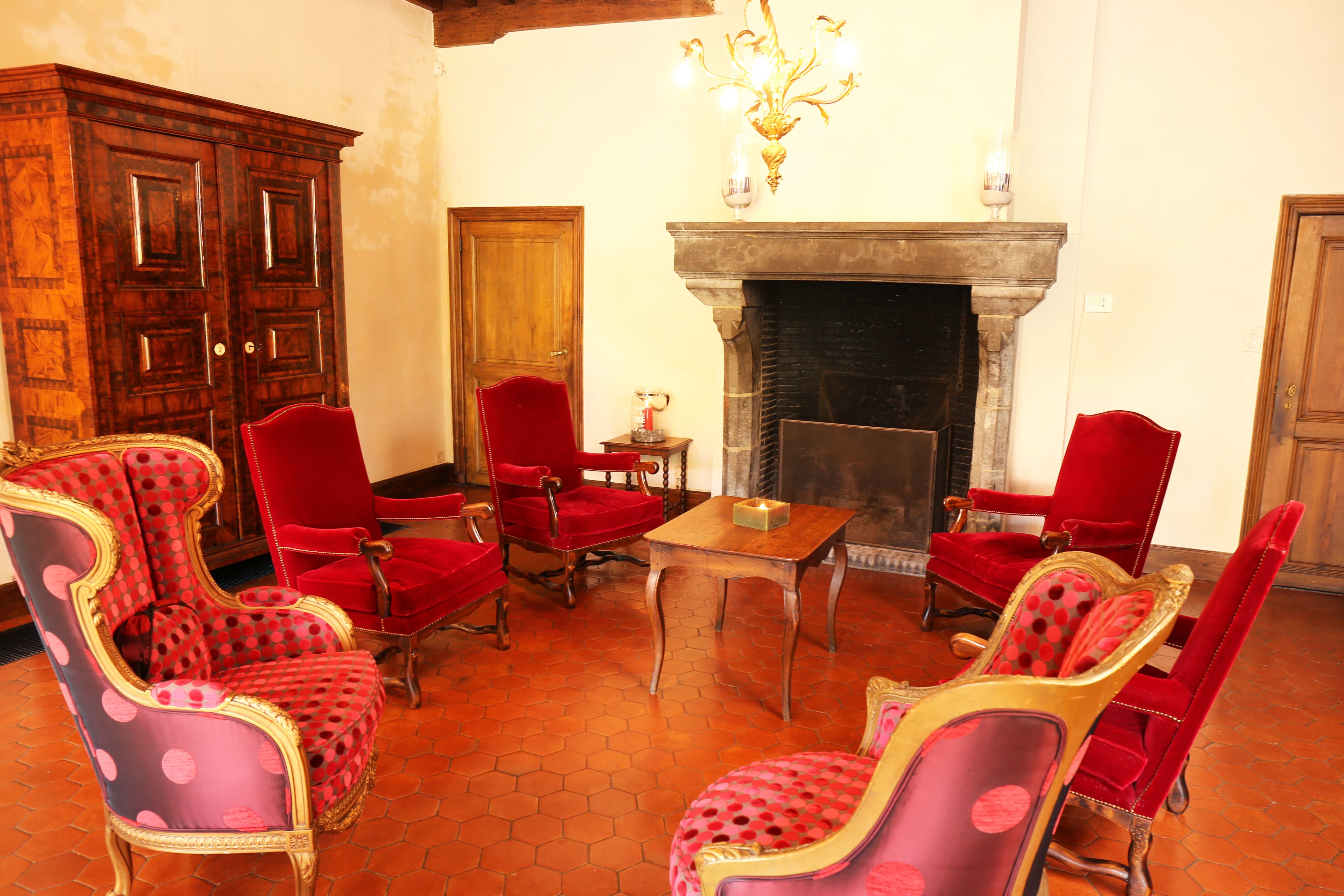 Um quarto com lareira, poltronas estofadas em vermelho, um armário de madeira e um candelabro.