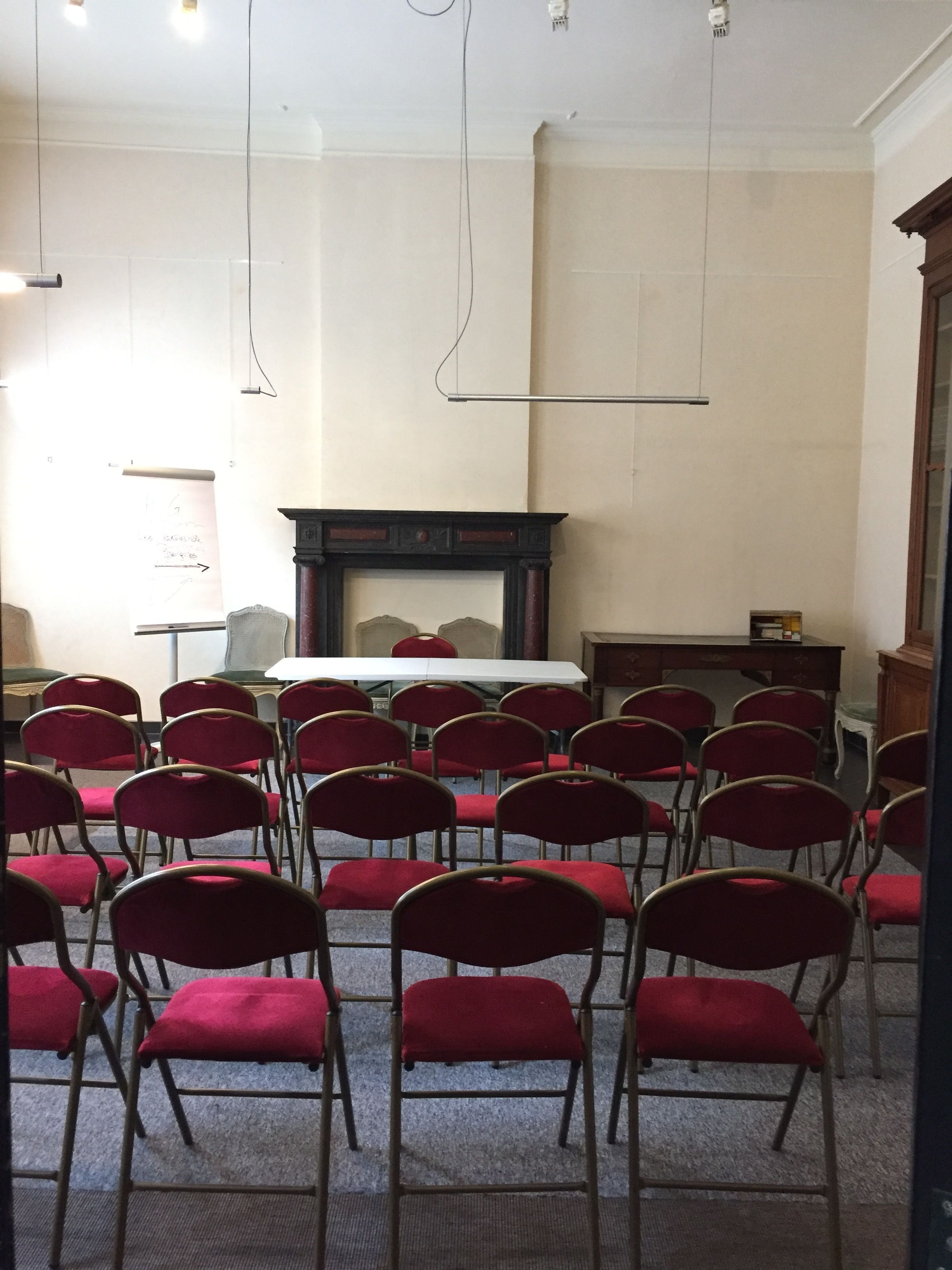 Une salle de conférence avec des rangées de chaises rouges, une table blanche, une cheminée et un tableau blanc.