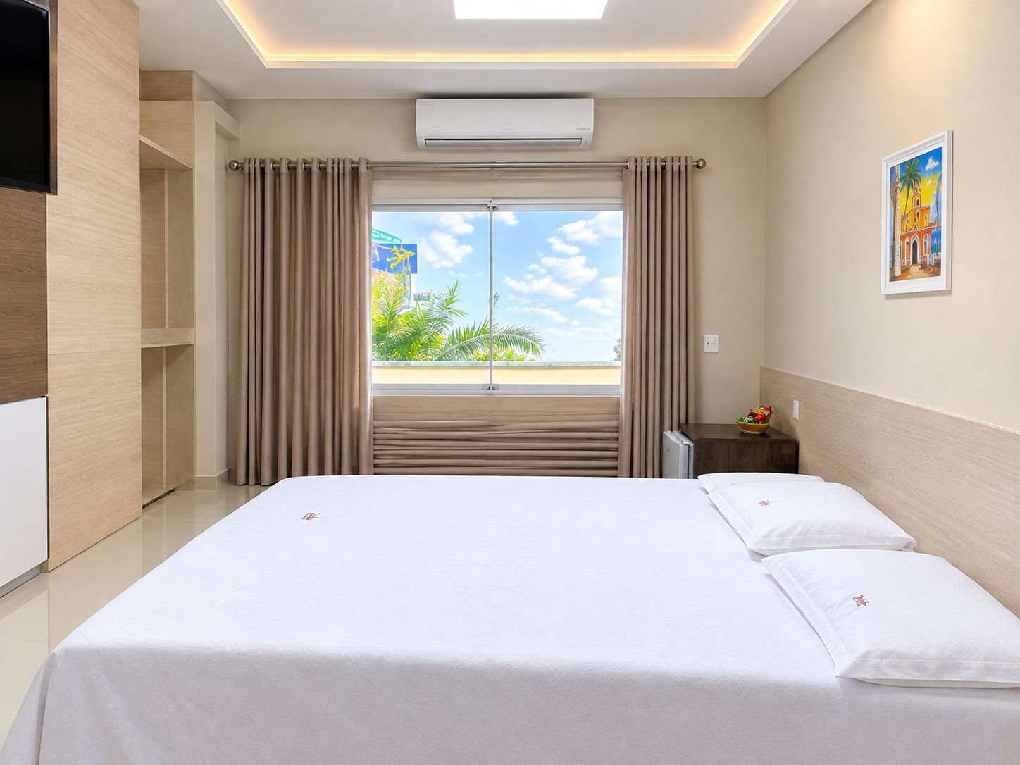 A hotel room with a bed, curtains, an air conditioning unit, a nightstand, a painting, and a view of palm trees.