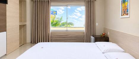 A hotel room with a bed, curtains, an air conditioning unit, a nightstand, a painting, and a view of palm trees.