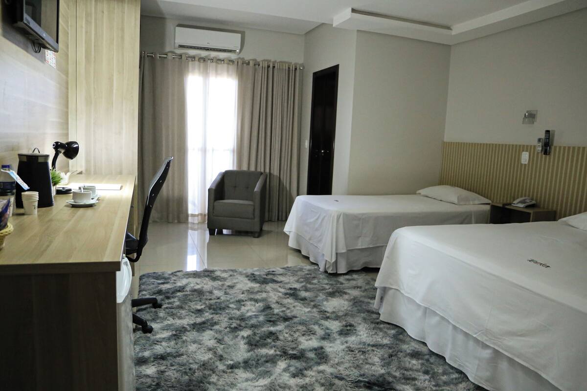 A hotel room with two beds, a desk with a chair, a small table with a lamp, and a window with curtains.