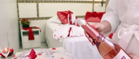 A person in a white robe pouring champagne into a glass. The setting appears to be a hotel room with a bed covered in white and red linens, a bedside table with a red cloth, and a mirror on the wall.