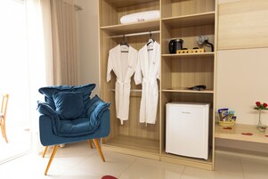 Quarto moderno com poltrona azul, guarda-roupa de madeira com roupões, frigorífico branco e pequena mesa com um vaso de flores.