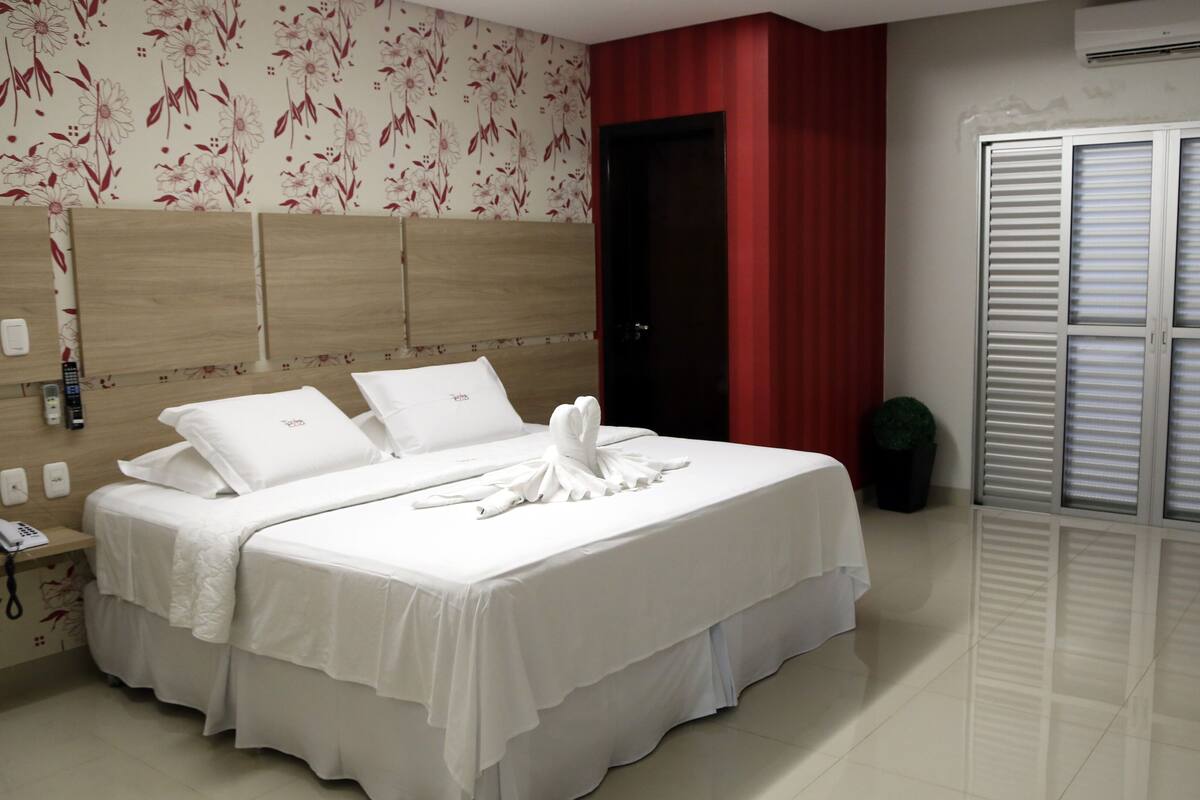A hotel room with a bed, a bedside table with a phone, a wall-mounted air conditioner, and a window with blinds.