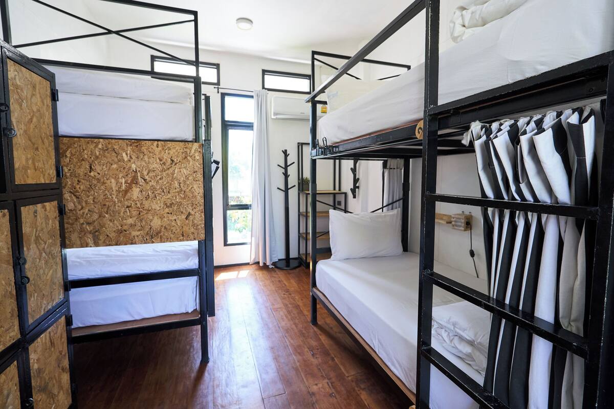 A dormitory-style room with bunk beds, wooden flooring, and a window with curtains.