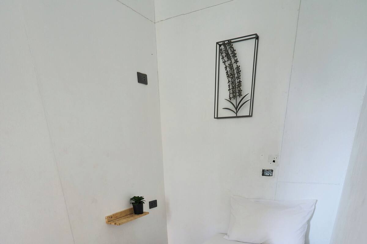 A neatly made bed with a white pillow and a decorative flower arrangement, a small wooden shelf with a plant, and a wall-mounted art piece.