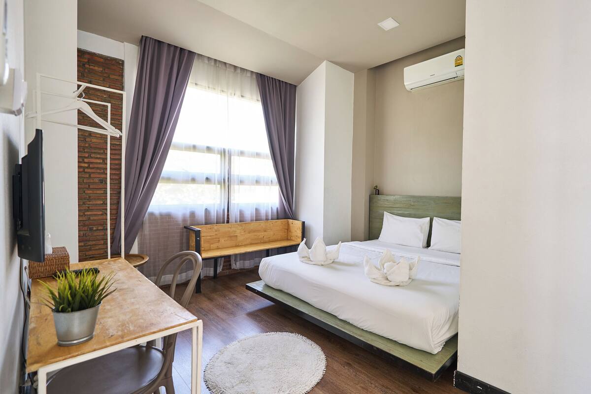 A hotel room with a bed, a desk with a plant, a bench, and a TV.