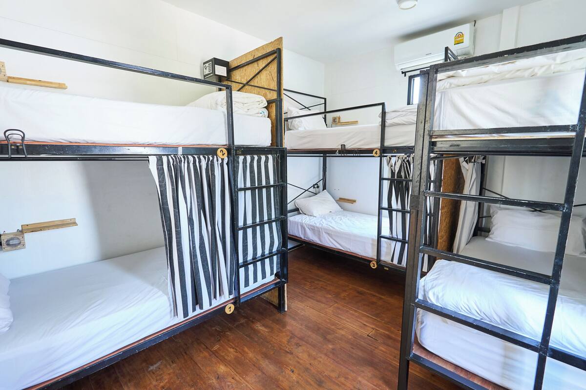 A dormitory room with bunk beds, wooden flooring, and a white wall.
