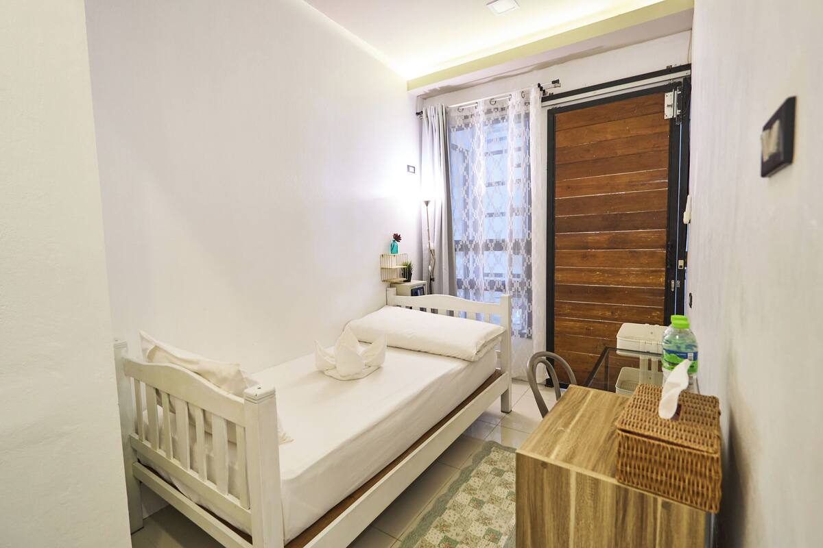 A small, clean room with a single bed, a wooden door, a small table with a bottle, and a chair.