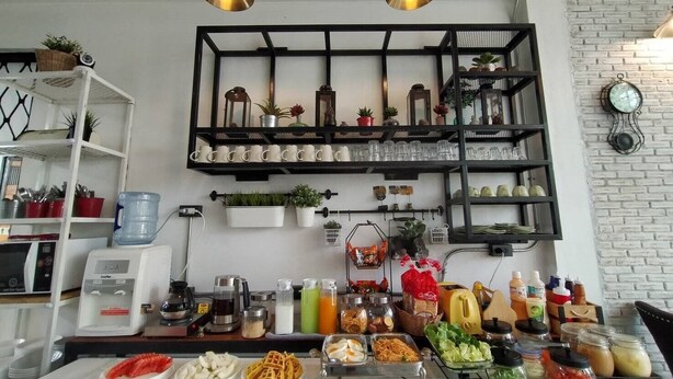 A kitchen area with a counter displaying food items, shelves with kitchenware, and hanging plants. - Sherloft Home & Hostel (Chiang Mai)
