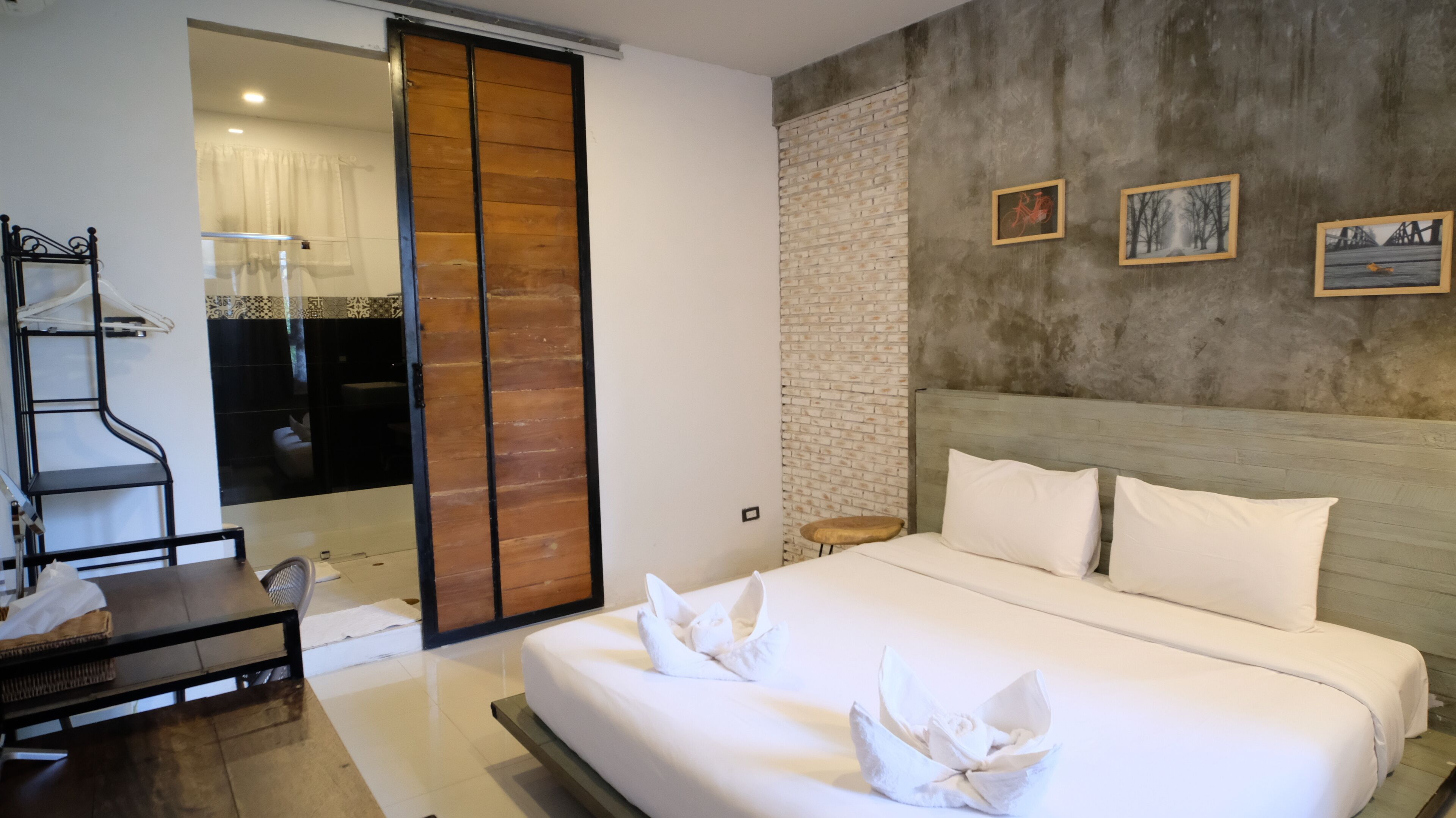 A modern hotel room with a large bed, a wooden wardrobe, a desk, and a bathroom with a glass door.