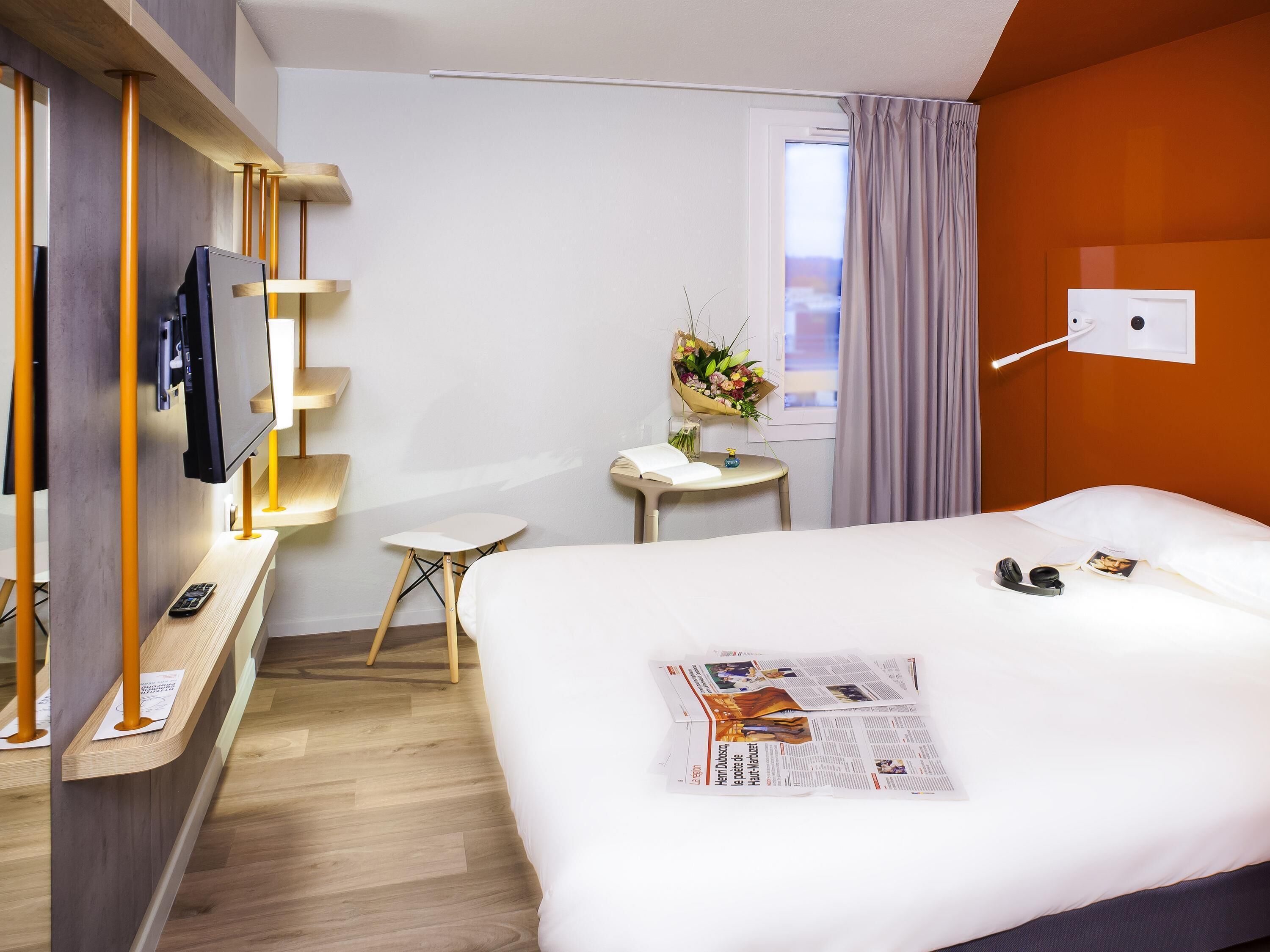 Ein Hotelzimmer mit Bett, Schreibtisch, Fernseher, einem Fenster mit Vorhängen und einer Wandleuchte.