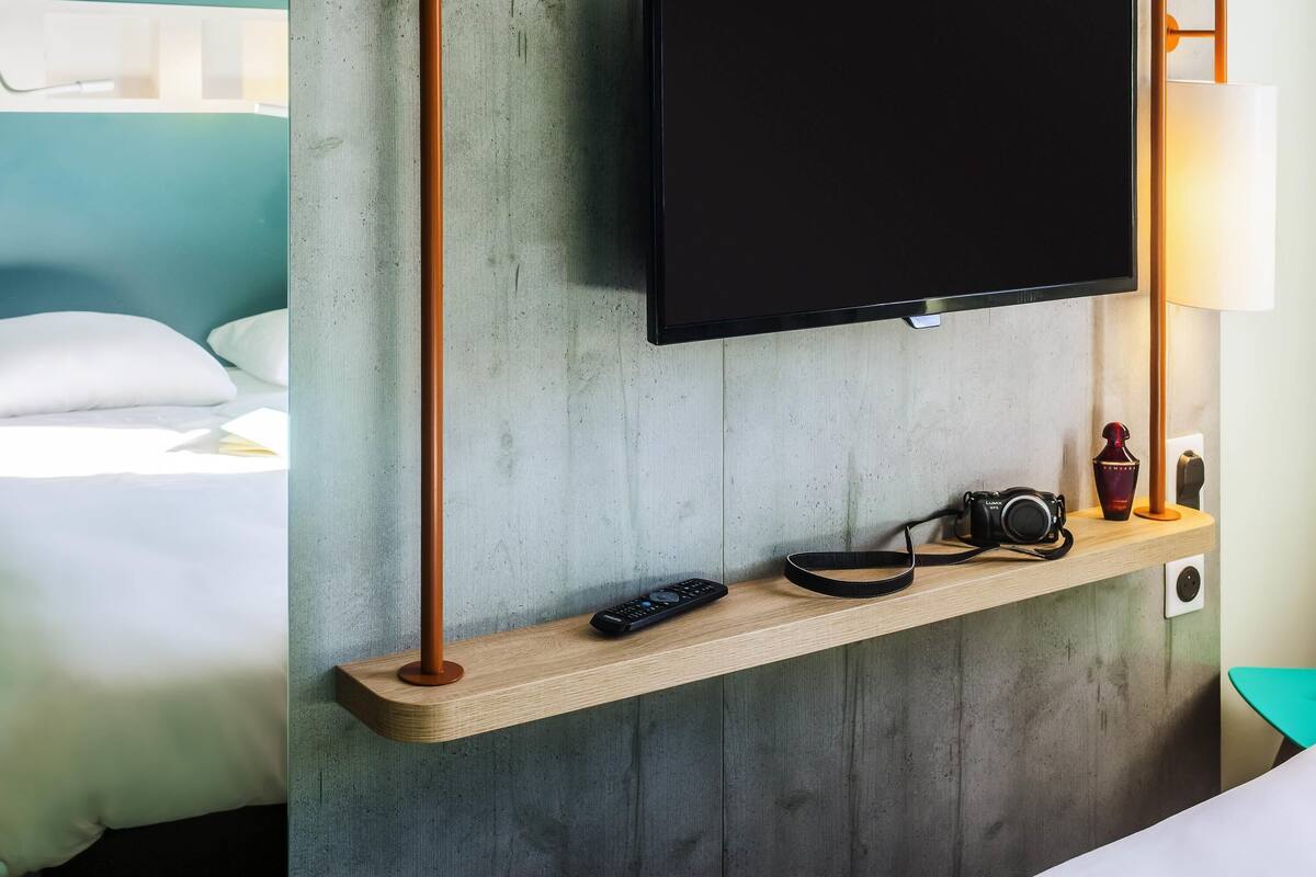 Habitación de hotel moderna con televisor montado, repisa de madera con una cámara y control remoto, y una lámpara de noche.