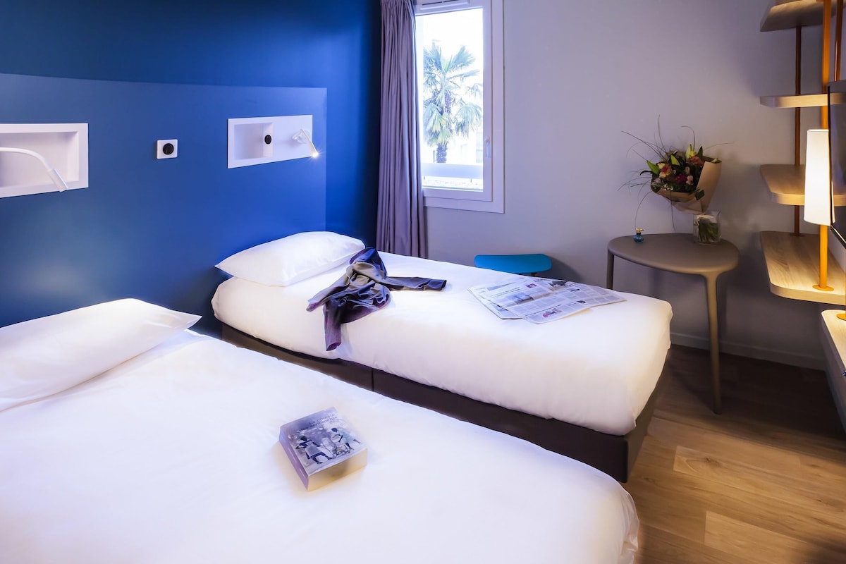 Une chambre d’hôtel avec deux lits, une petite table, une lampe et une fenêtre donnant sur des palmiers.