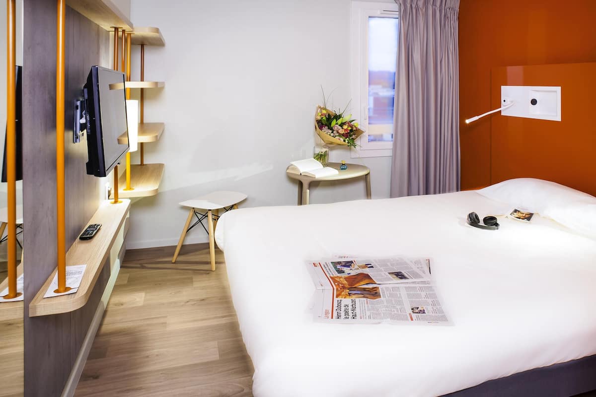 Ein Hotelzimmer mit Bett, Schreibtisch, Fernseher, einem Fenster mit Vorhängen und einer Wandleuchte.