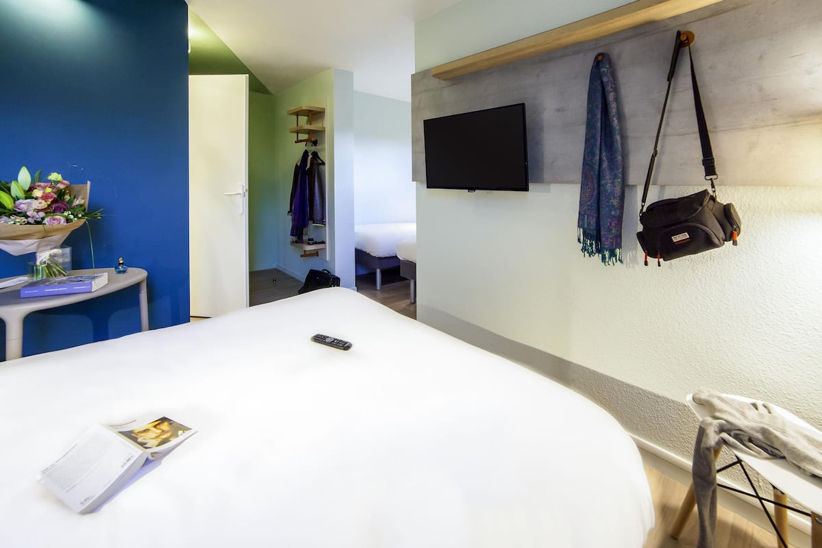 Habitación de hotel con cama, televisor montado en la pared, mesita de noche con control remoto, pequeña mesa con un libro y un jarrón de flores, y perchero con chaqueta y bufanda.