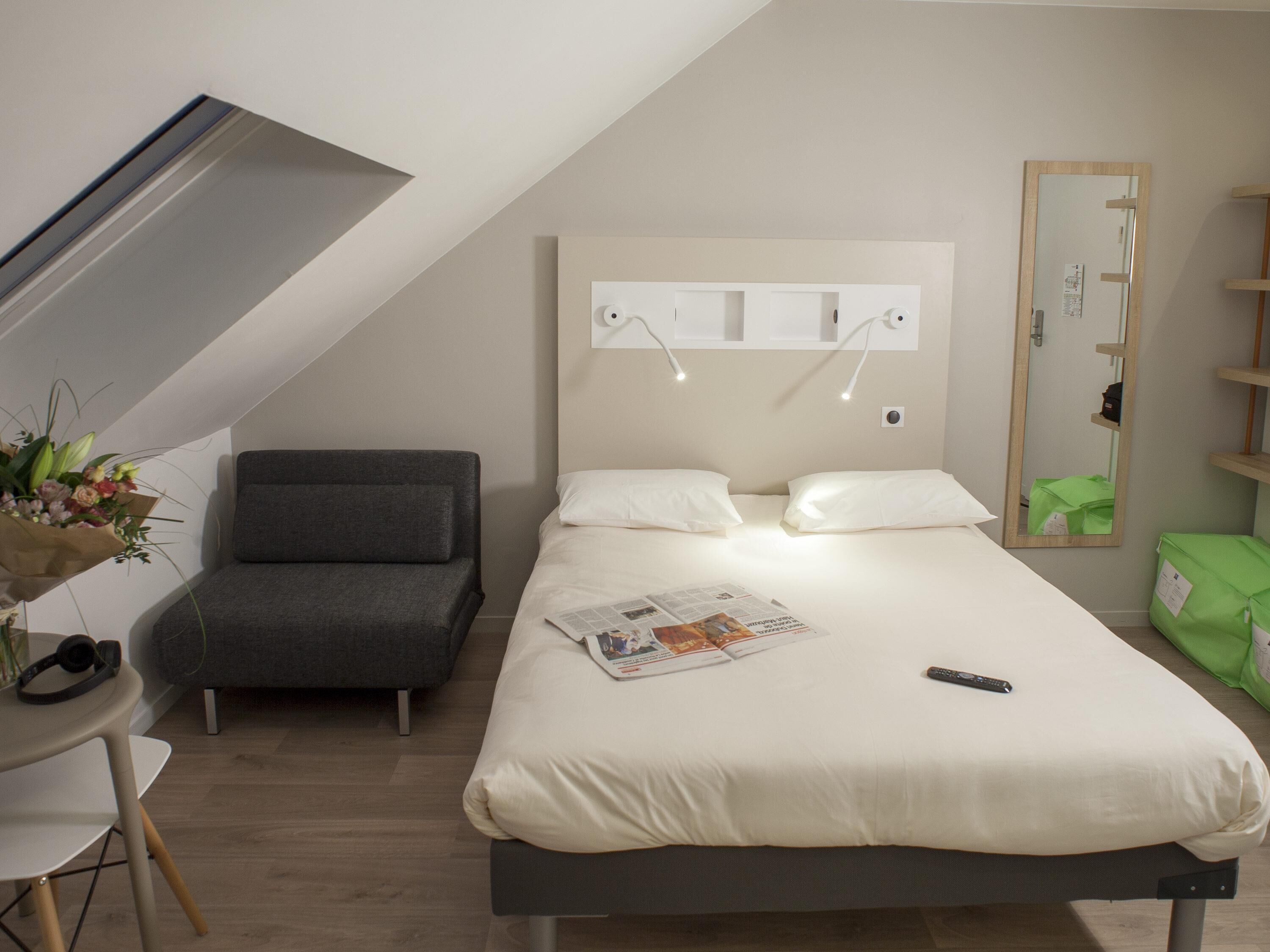 A modern hotel room with a bed, a small desk, a chair, and a mirror.