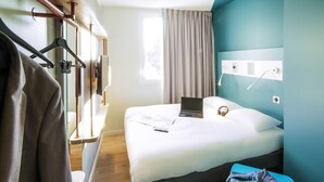 A hotel room with a bed, a desk, a chair, a coat rack, and a window with curtains.