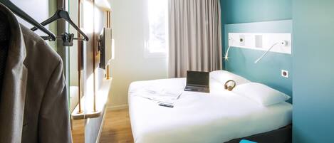 Hotellihuone, jossa on sänky, työpöytä, tuoli, naulakko ja ikkuna, jossa on verhot.
