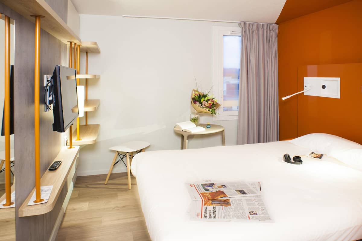 Een hotelkamer met een bed, een televisie, een klein tafeltje met bloemen en een raam met gordijnen.