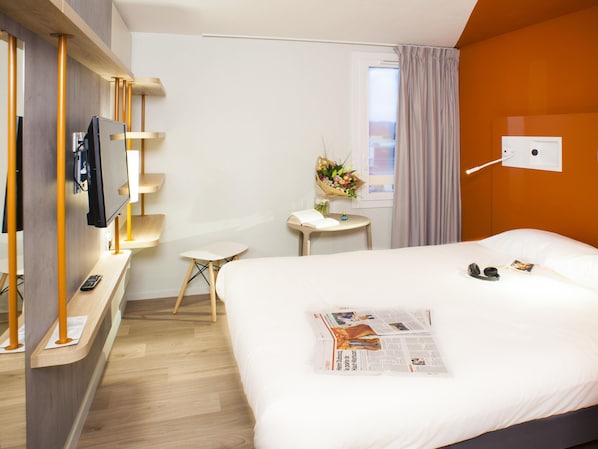Une chambre d’hôtel avec un lit, une télévision, une petite table agrémentée d’un arrangement floral et une fenêtre avec des rideaux.