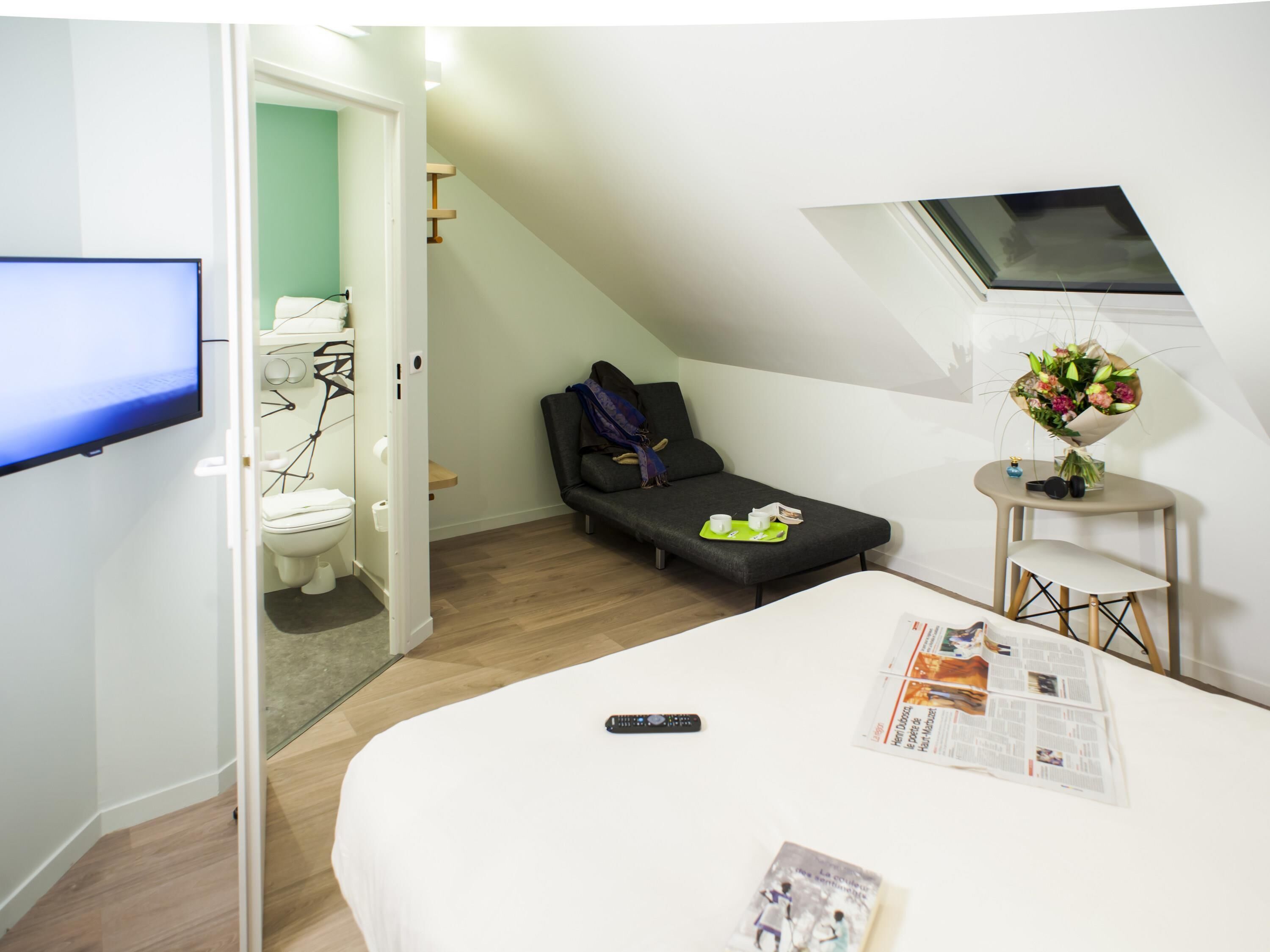 A compact room with a bed, a small bedside table with a newspaper, a remote control, and a window with a view of the sky.