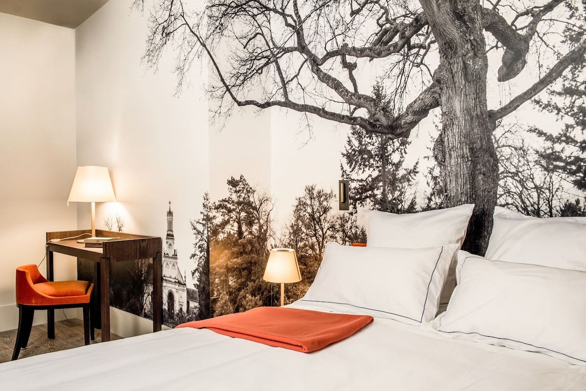 A hotel room with a bed, two bedside lamps, a desk with a chair, and a large tree mural on the wall.