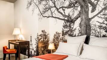 A hotel room with a bed, two bedside lamps, a desk with a chair, and a large tree mural on the wall.