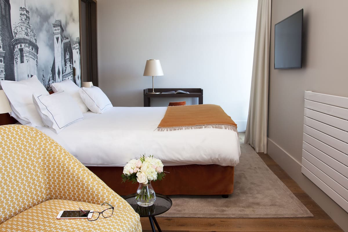 A hotel room with a bed, a nightstand, a lamp, a TV, a sofa, and a vase with flowers.