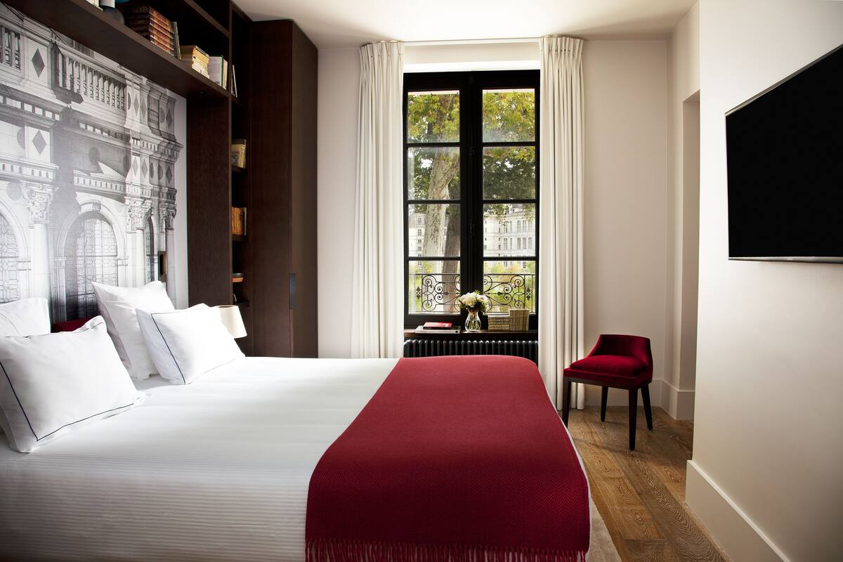 A hotel room with a large bed, a red throw blanket, a red chair, a bookshelf, and a large window with a view of buildings.