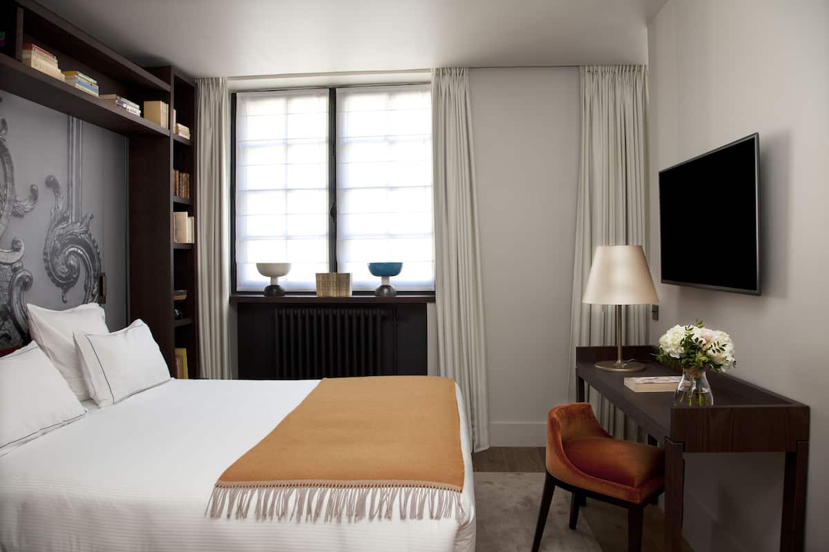 A hotel room with a large bed, a desk with a lamp, a TV, and a bookshelf.