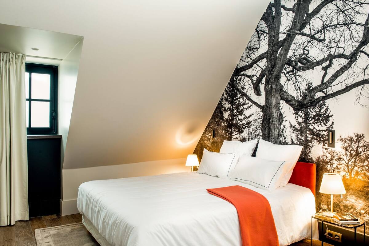 A bedroom with a bed, a nightstand, a lamp, a window with curtains, and a wall mural.