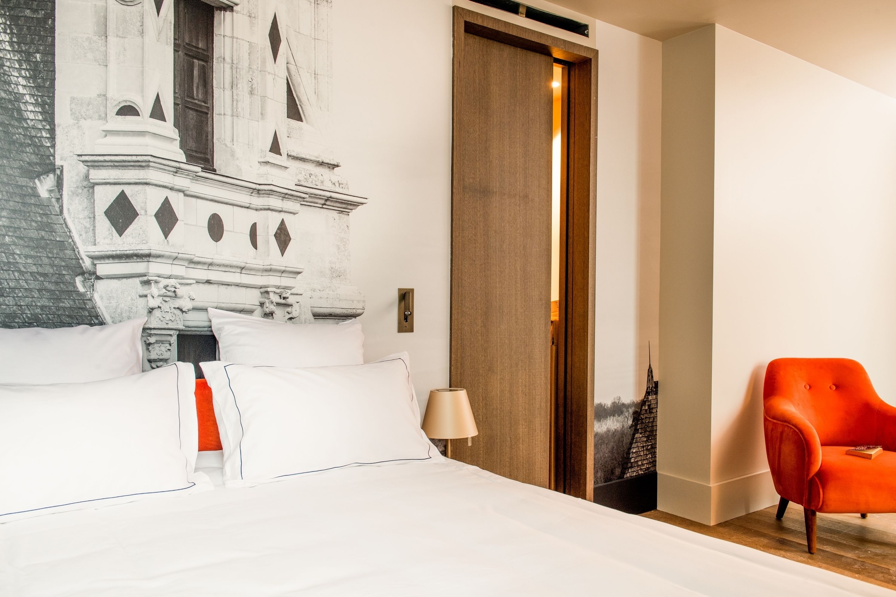 Quarto de hotel com cama, mesa de cabeceira, luminária, poltrona vermelha e uma grande pintura na parede.