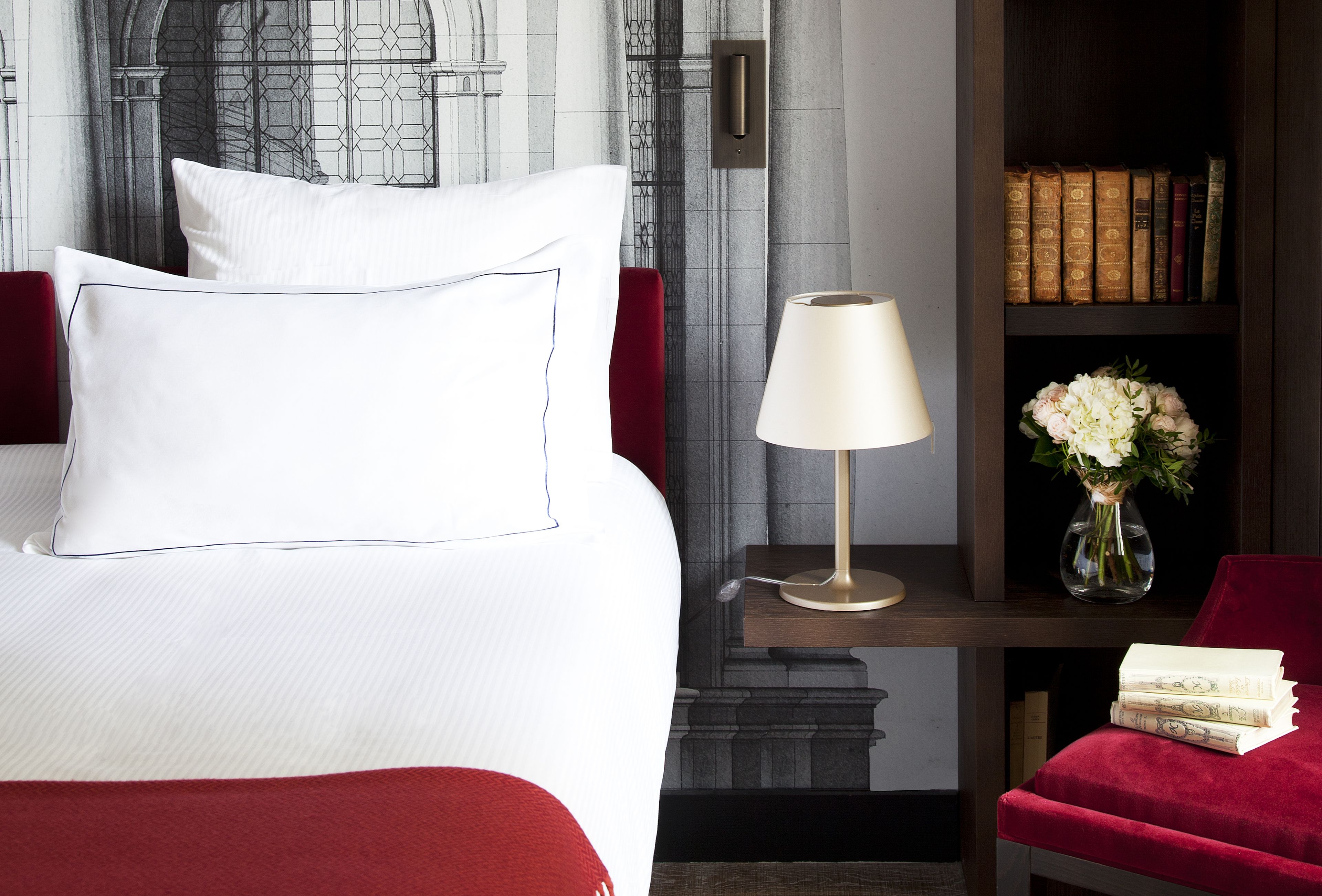 Una habitación de hotel con una cama, una mesita de noche con lámpara, un jarrón con flores, una estantería con libros y una silla roja.