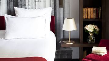 Una habitación de hotel con una cama, una mesita de noche con lámpara, un jarrón con flores, una estantería con libros y una silla roja.