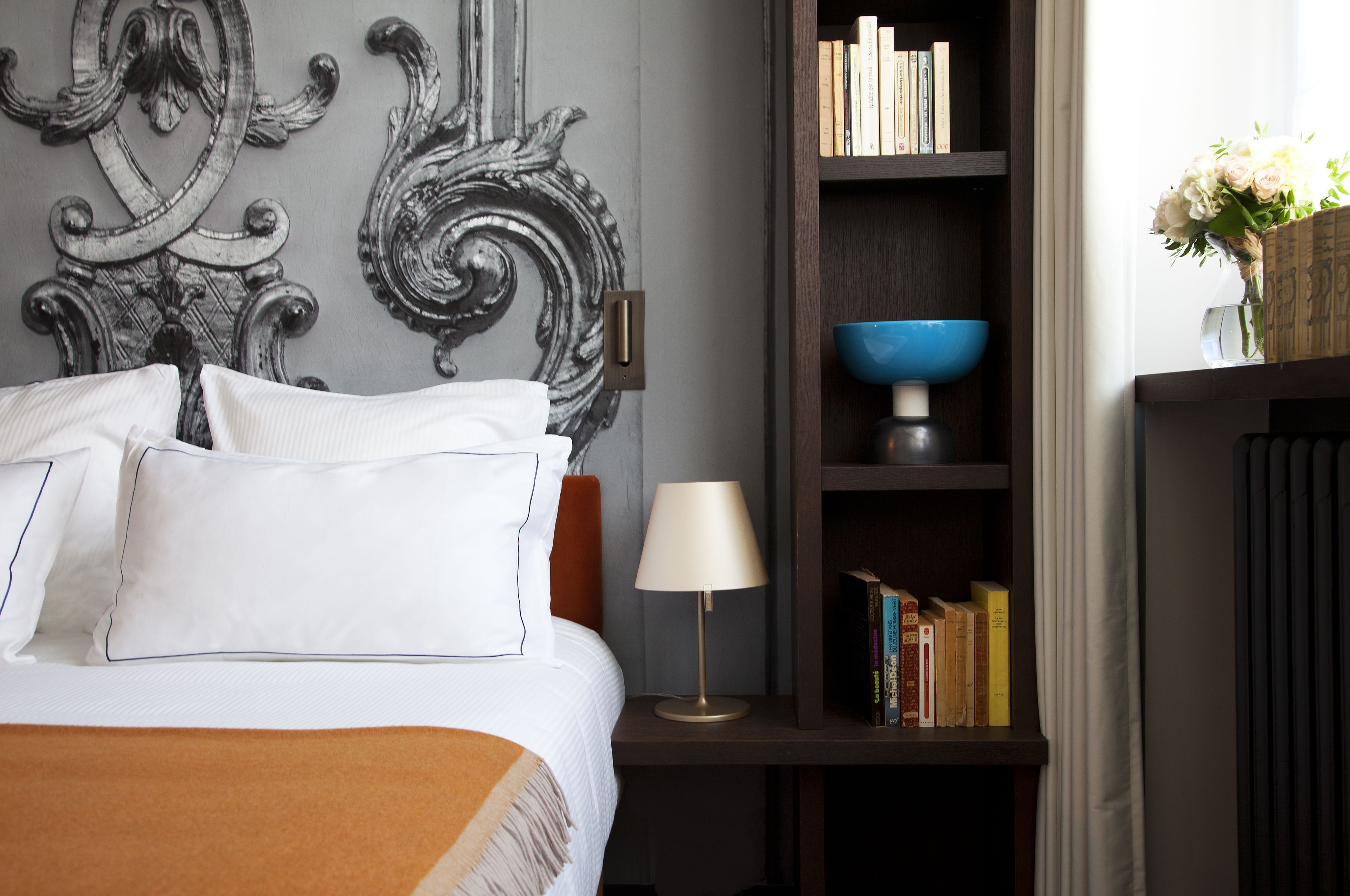 Una cama bien hecha con almohadas y una colcha, una mesita de noche con una lámpara y libros, una pared decorativa y una estantería con libros y un florero.
