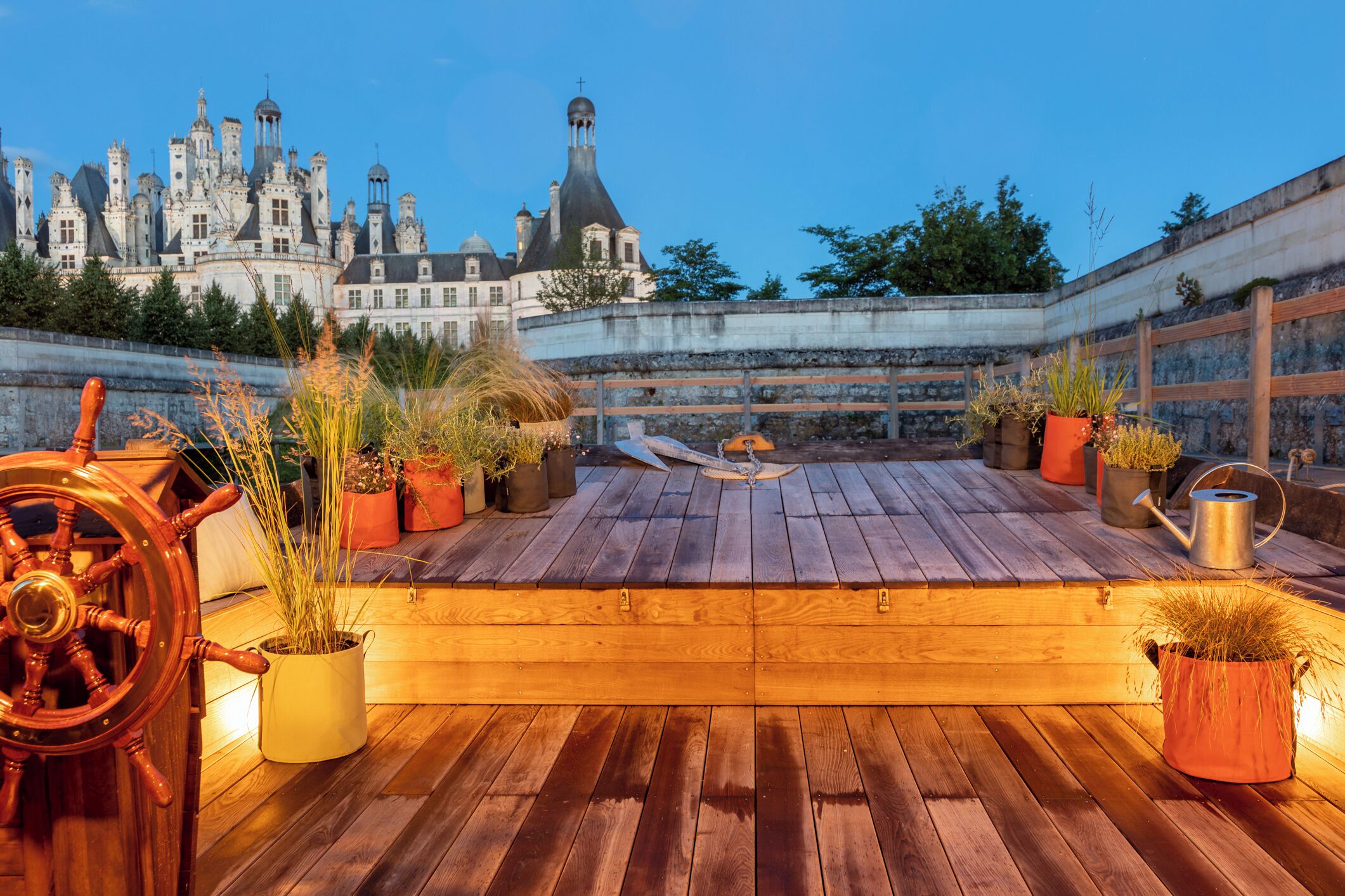 A wooden deck with potted plants and a ship's wheel, overlooking a historic castle.