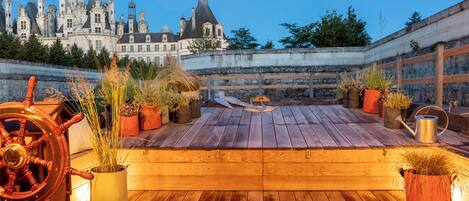 A wooden deck with potted plants and a ship's wheel, overlooking a historic castle.