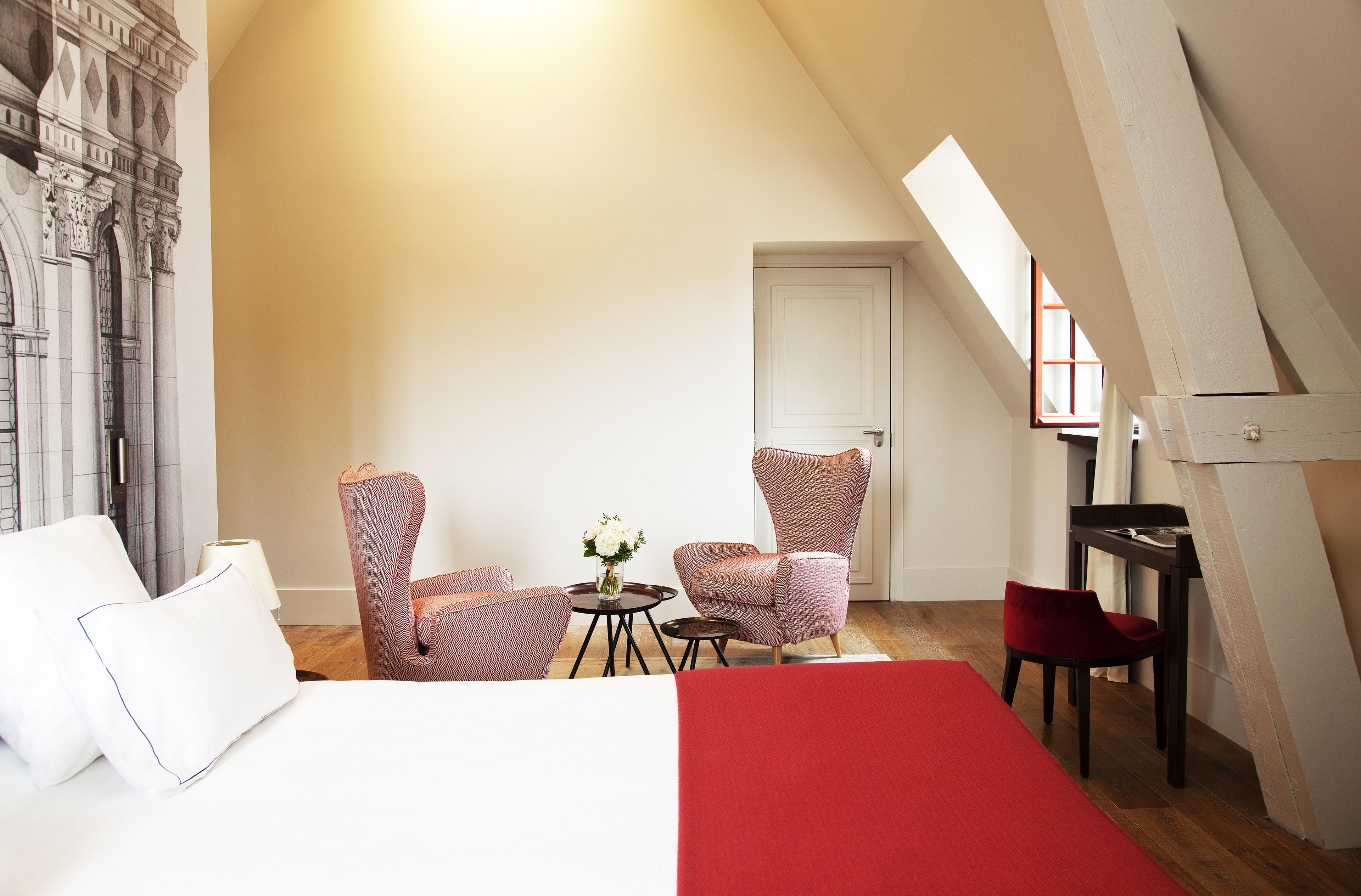 Une chambre à coucher avec un lit, deux chaises, une petite table sur laquelle se trouve un vase, et une porte.