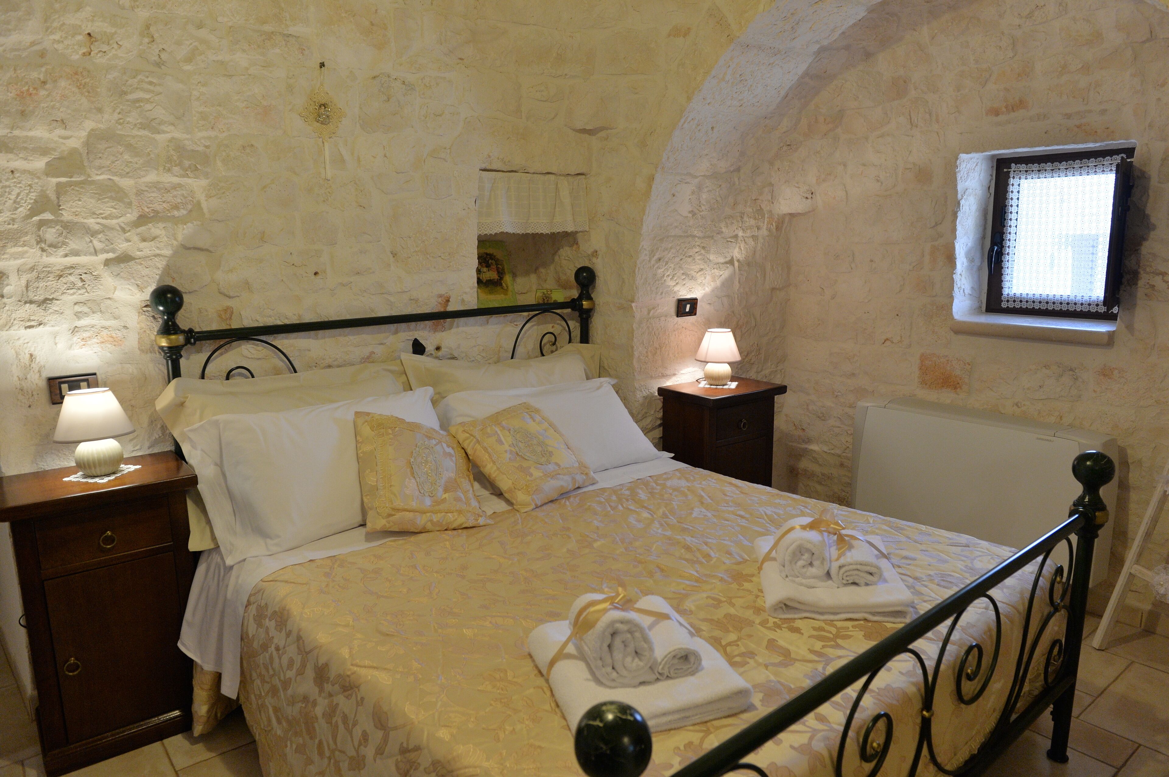 Une chambre avec un mur en pierre, un lit avec un cadre en métal, deux tables de chevet avec des lampes et une fenêtre donnant sur un beau paysage.
