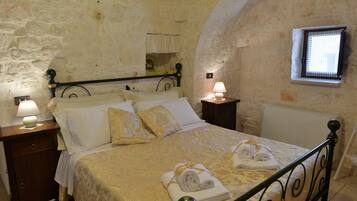 Une chambre avec un mur en pierre, un lit avec un cadre en métal, deux tables de chevet avec des lampes et une fenêtre donnant sur un beau paysage.