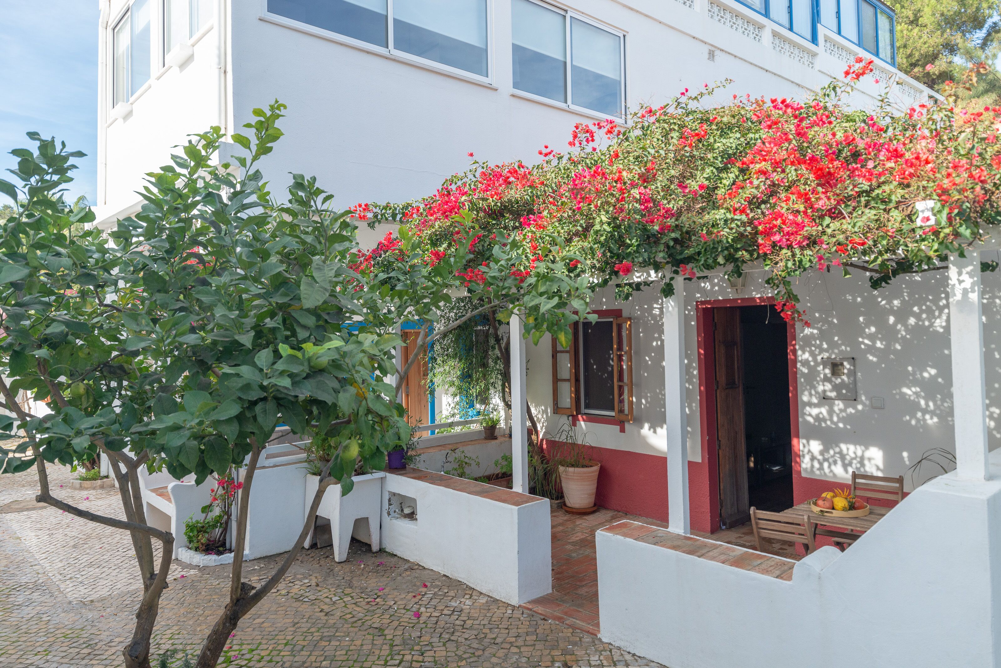 Un bâtiment blanc avec une porte rouge, un balcon orné de fleurs rouges et une cour dallée.