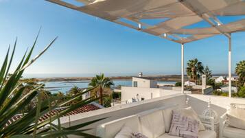Une terrasse avec un canapé et un fauteuil blancs, offrant une vue sur la côte, avec des palmiers et des bâtiments.