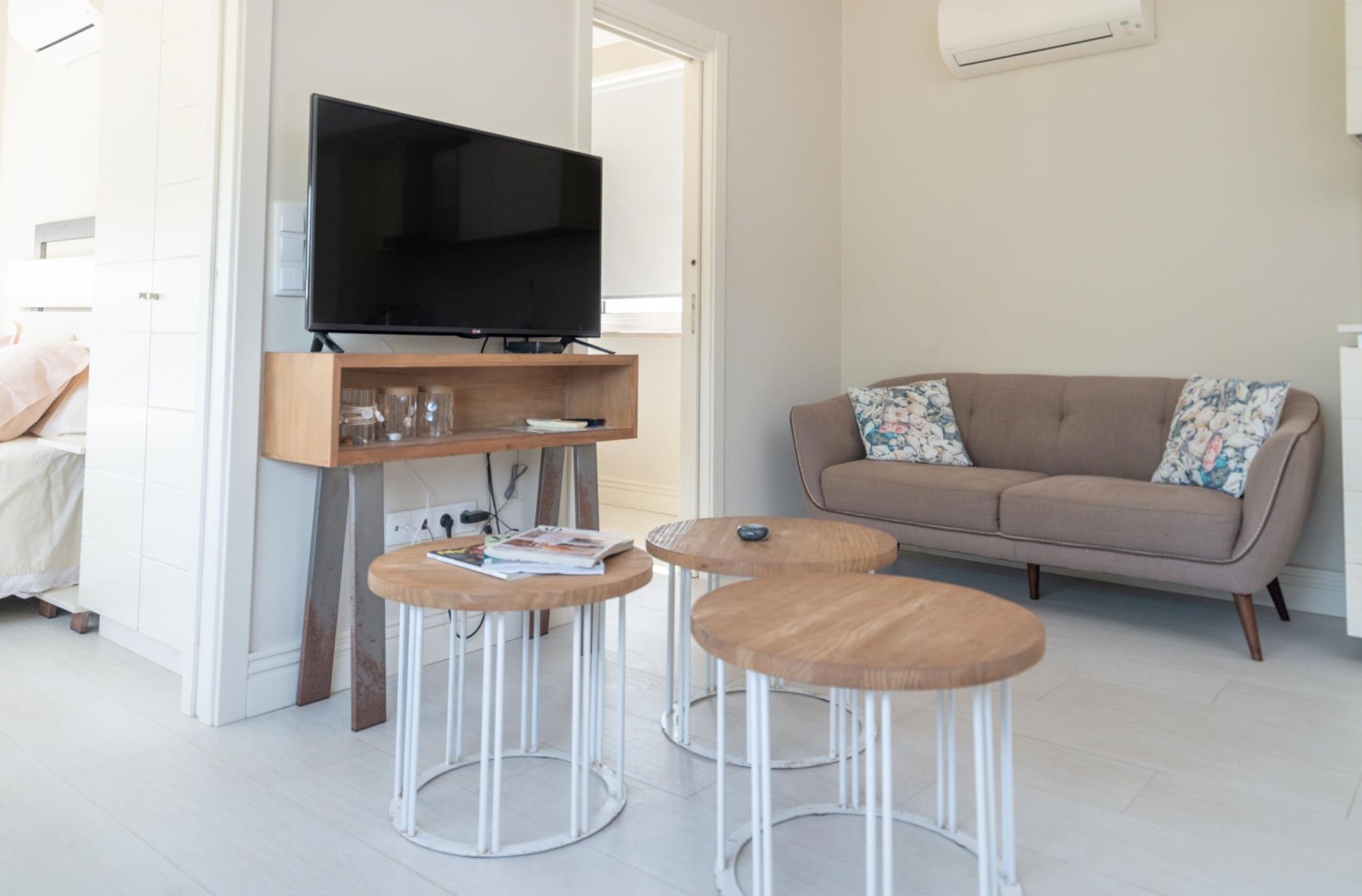 A modern living room with a flat-screen TV on a wooden stand, a beige sofa with floral cushions, and two round wooden tables.