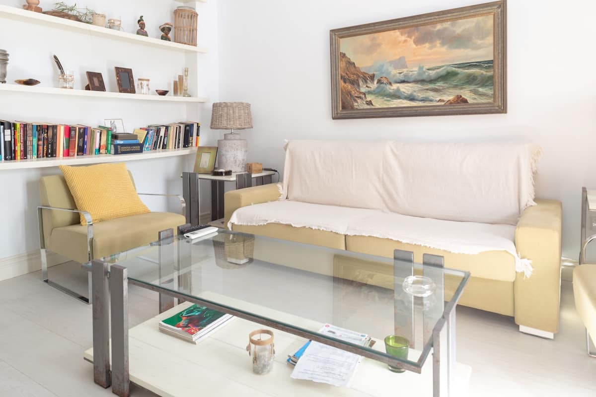 A living room with a glass coffee table, beige sofa, yellow cushion, and a bookshelf filled with books and decorative items.