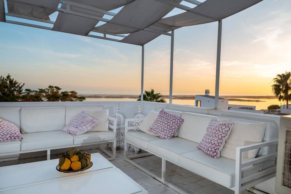 A white outdoor seating area with a view of the sea at sunset.
