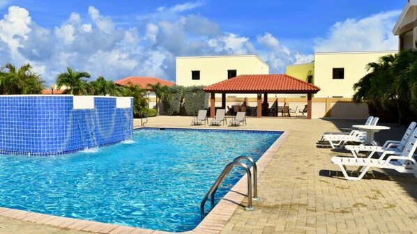 Una piscina con borde de azulejos, sillas de descanso y una zona de estar cubierta.