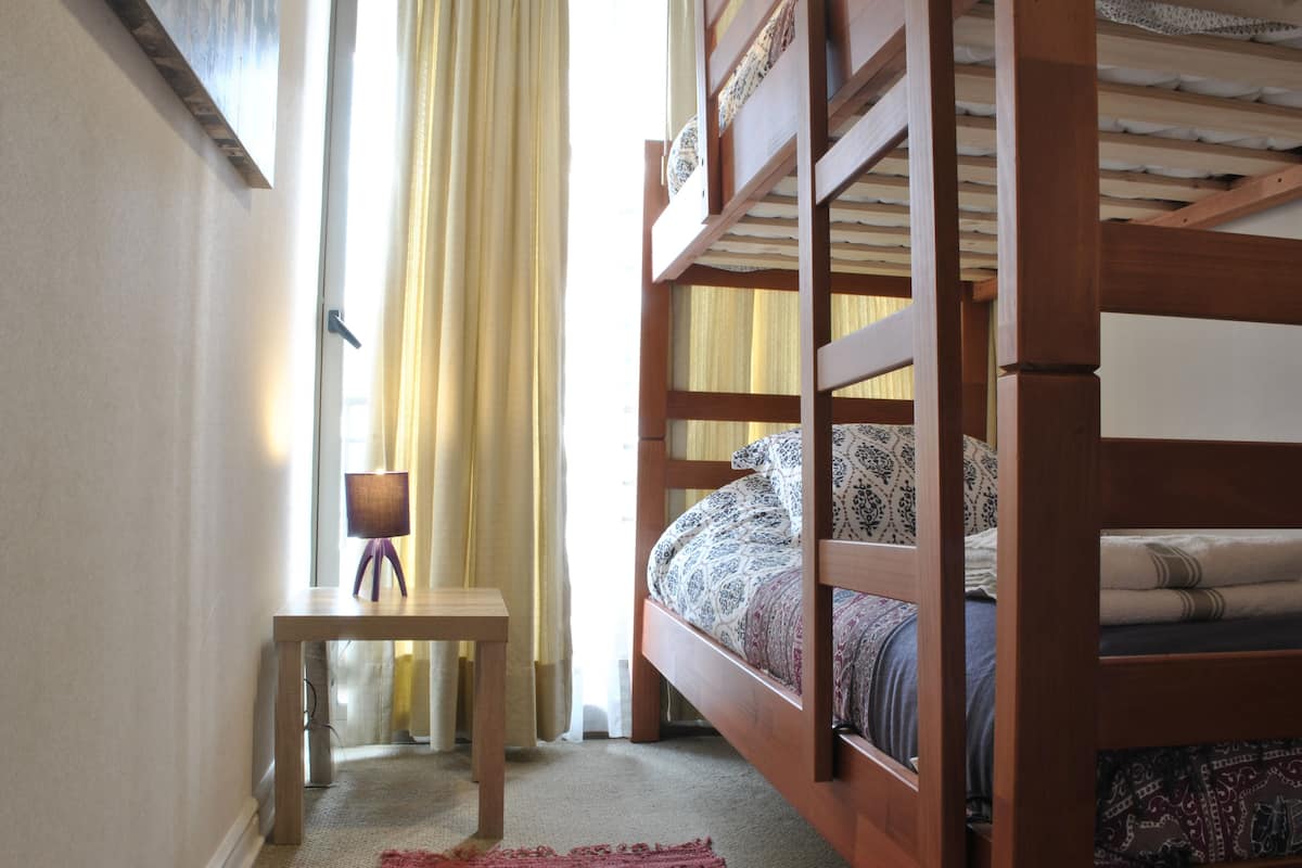 Ein Zimmer mit Etagenbett, einem kleinen Tisch mit Lampe und einem Fenster mit Vorhängen.